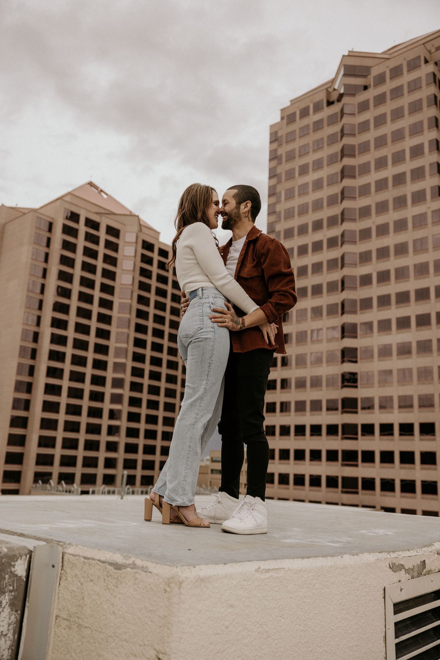 Couple sharing intimate moment on rooftop ledge with pink skyscrapers behind them.