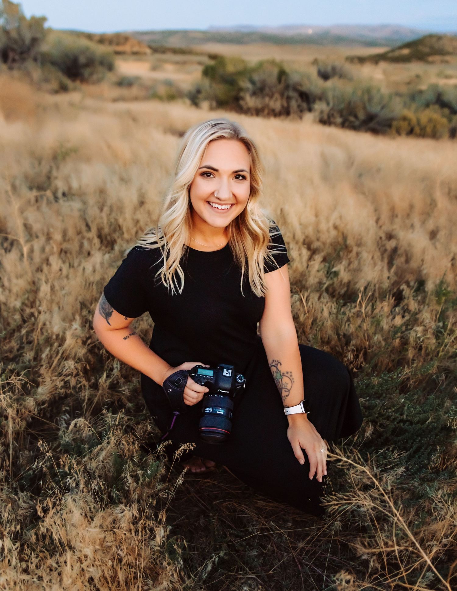 Professional photographer in black outfit poses with camera in golden desert landscape.