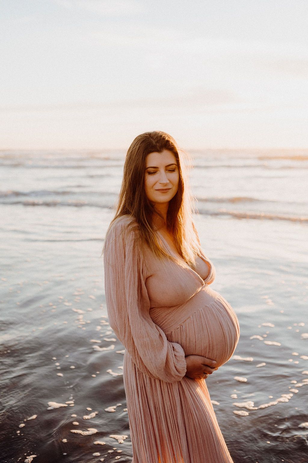 Maternity photoshoot on beach at sunset with flowing pink dress.