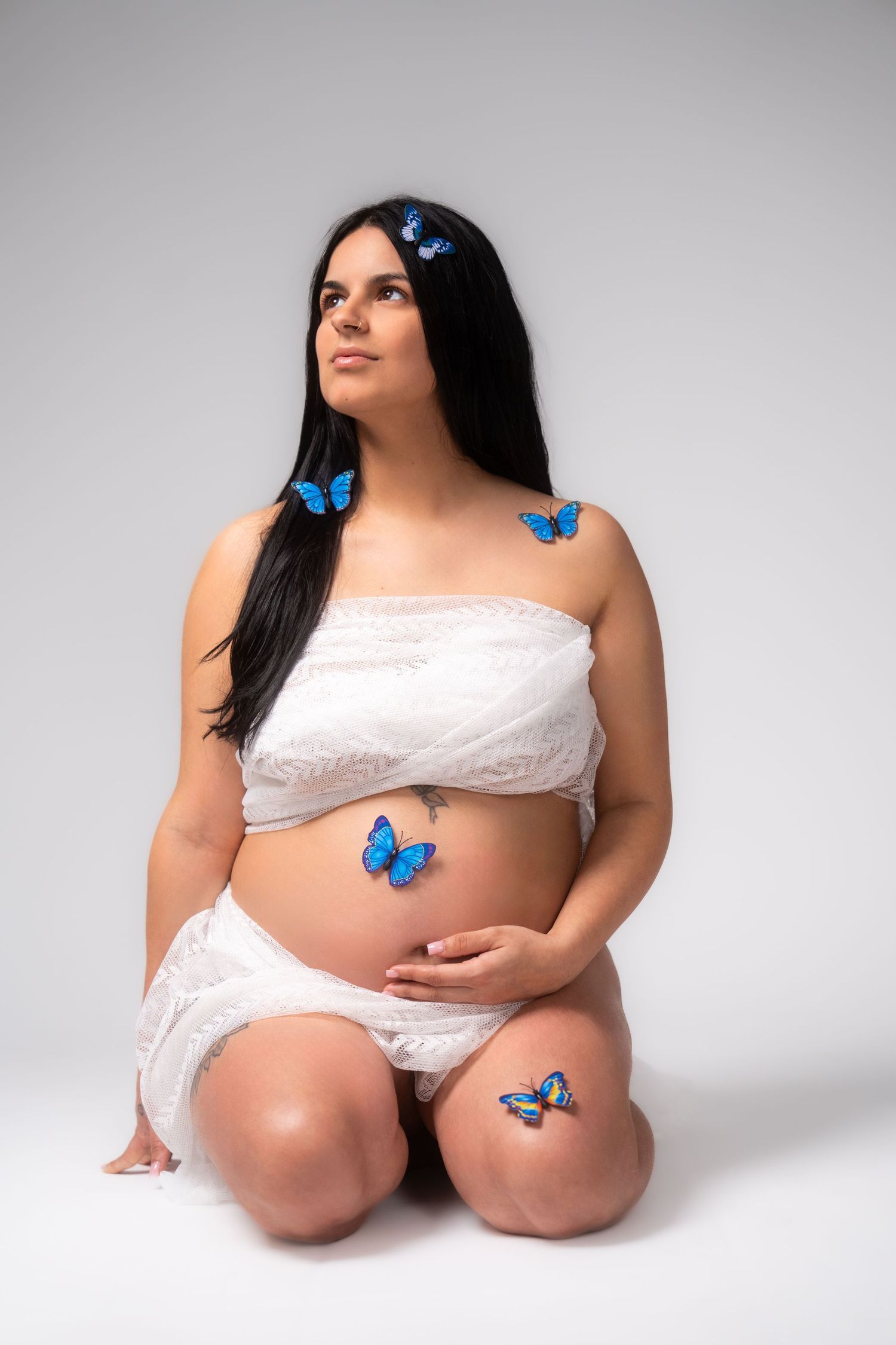 Expectant mother in white lace poses with decorative blue butterflies in studio setting.