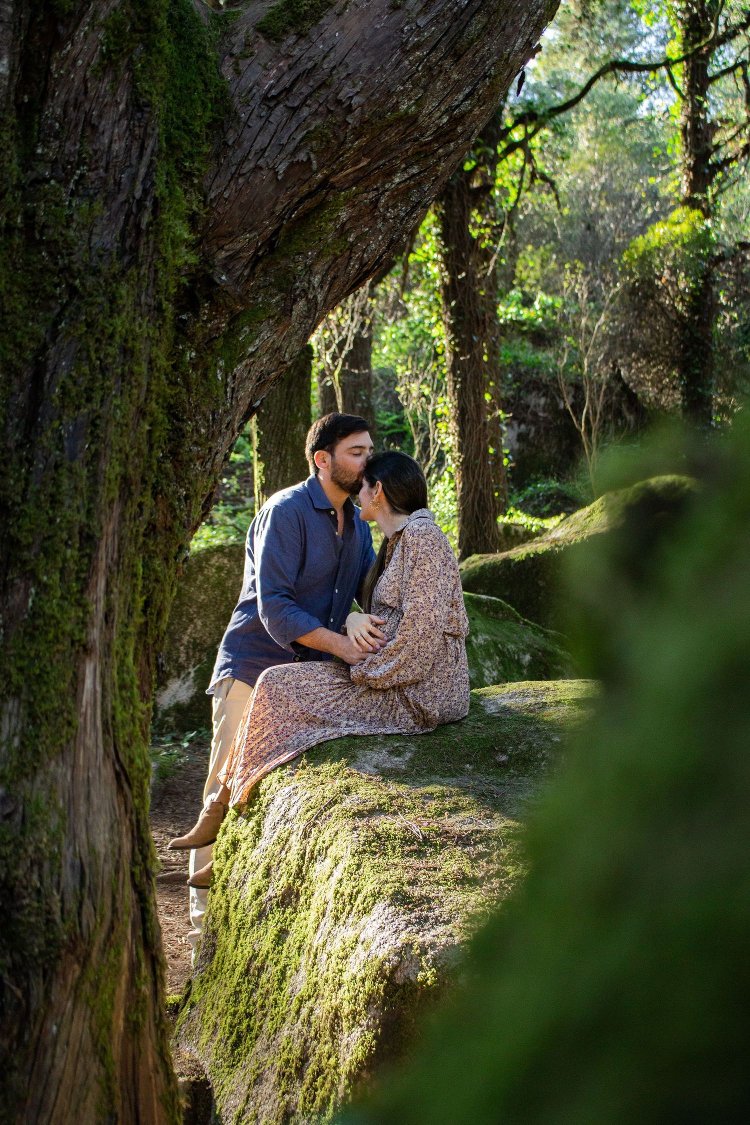 A romantic outdoor photoshoot with a couple sitting on a moss-covered rock surrounded by tall trees in a forest setting.