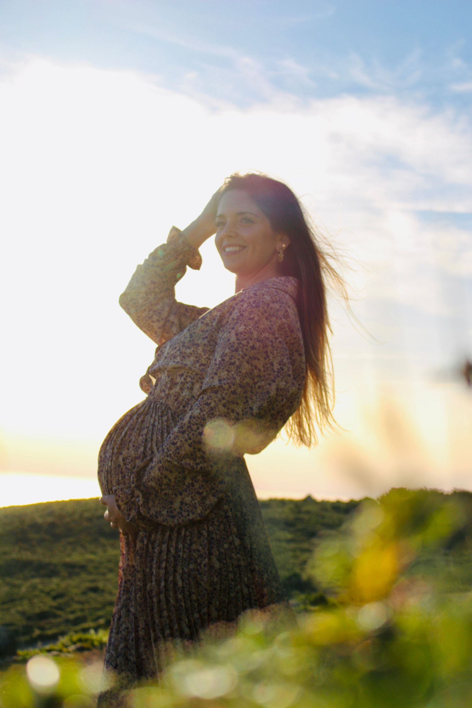 Silhouette of pregnant woman in flowing dress posing at sunset in nature setting.