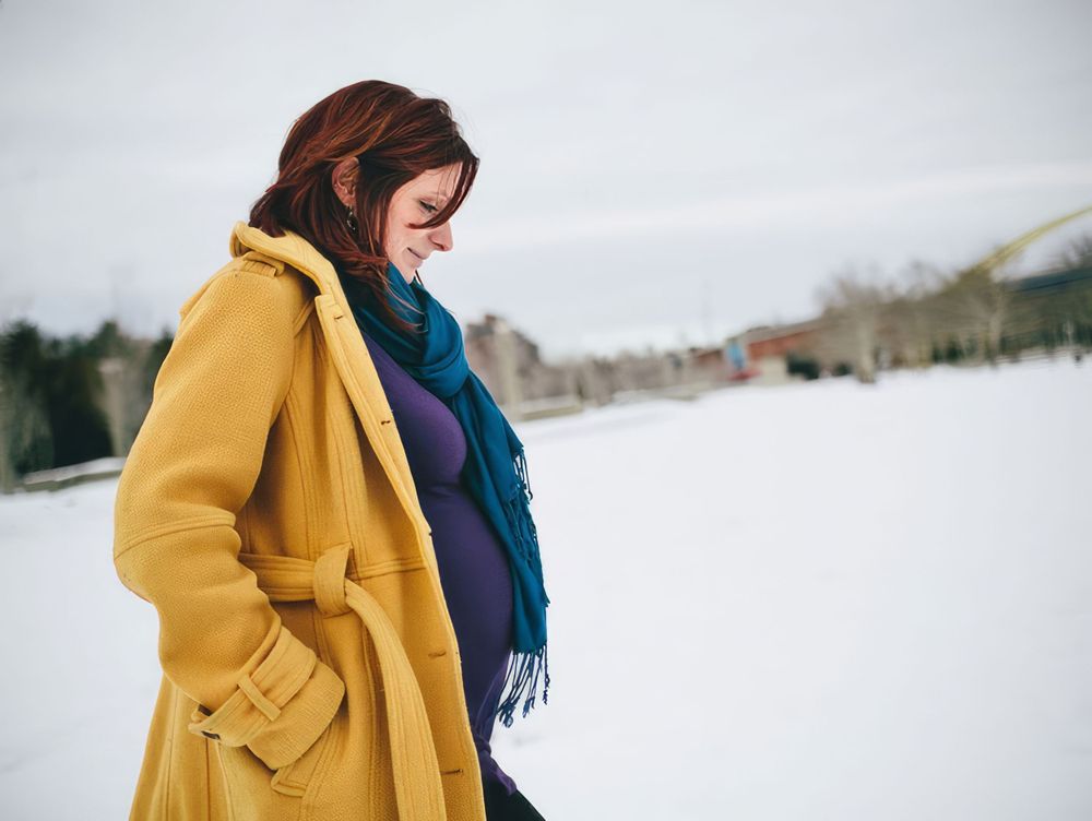 Pregnant woman wearing a yellow coat with a blue scarf walking in the snow.