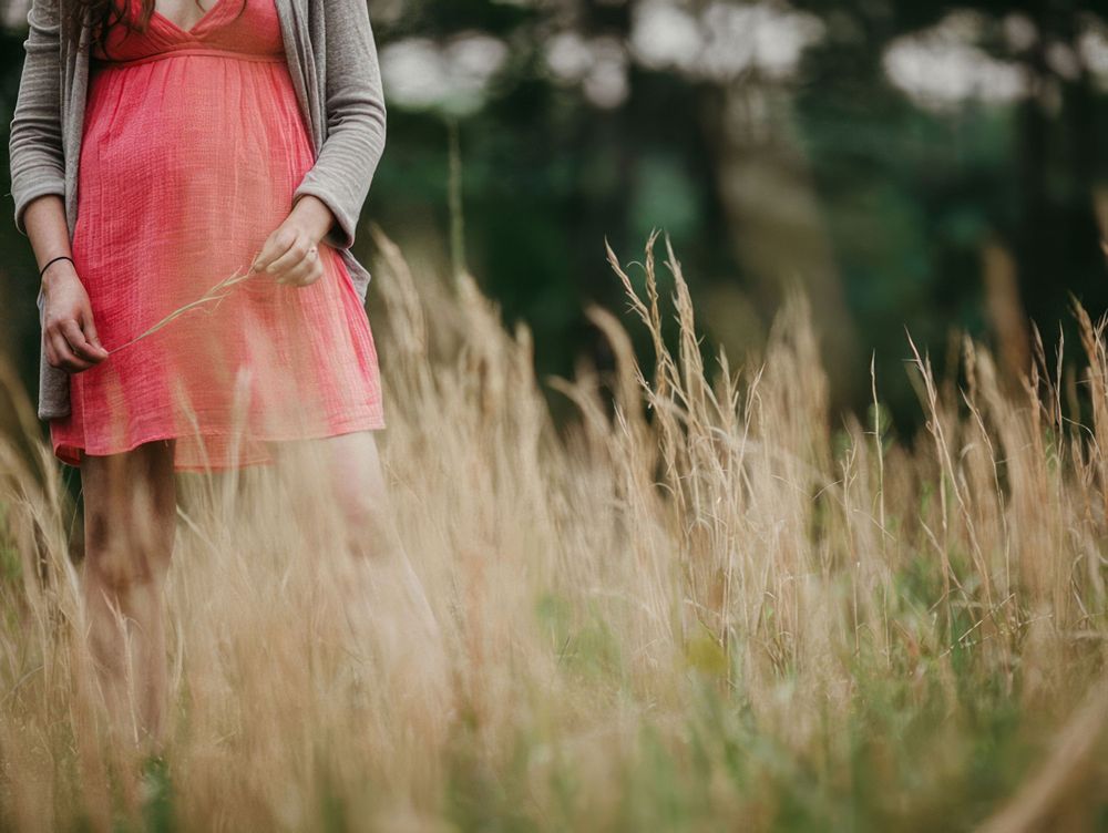 Professional maternity photograph of a woman wearing a pink dress and standing in the tall grass.