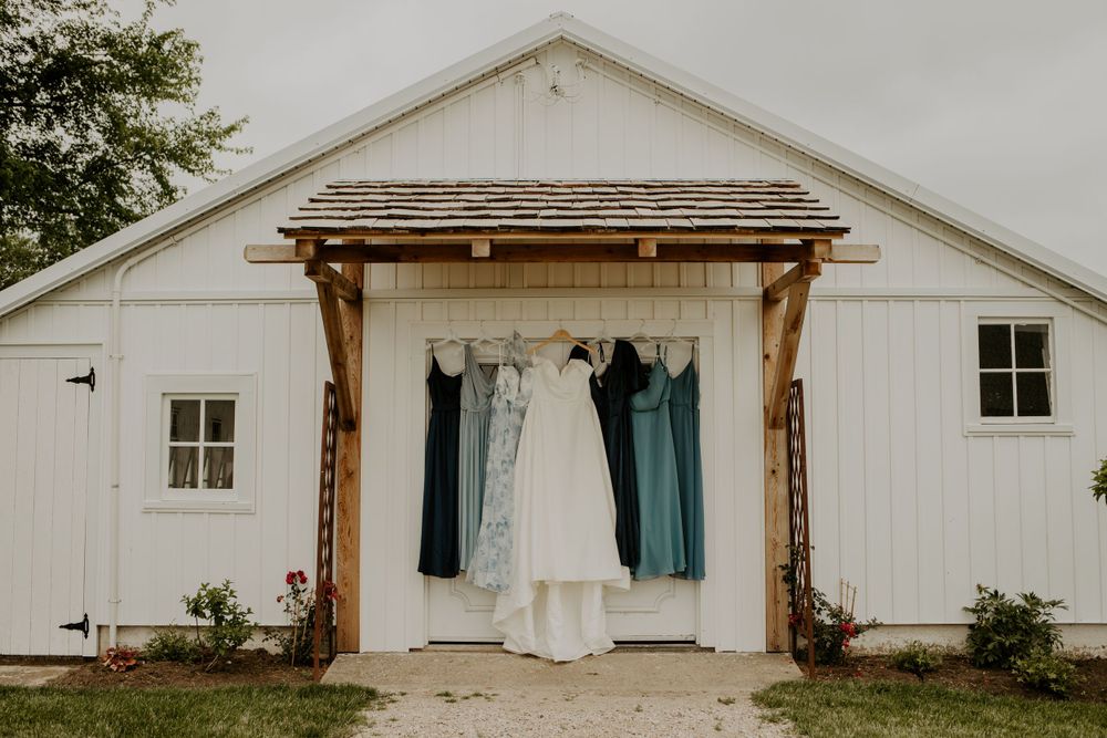 Wedding dresses and suits hang on display outside white barn building entrance.