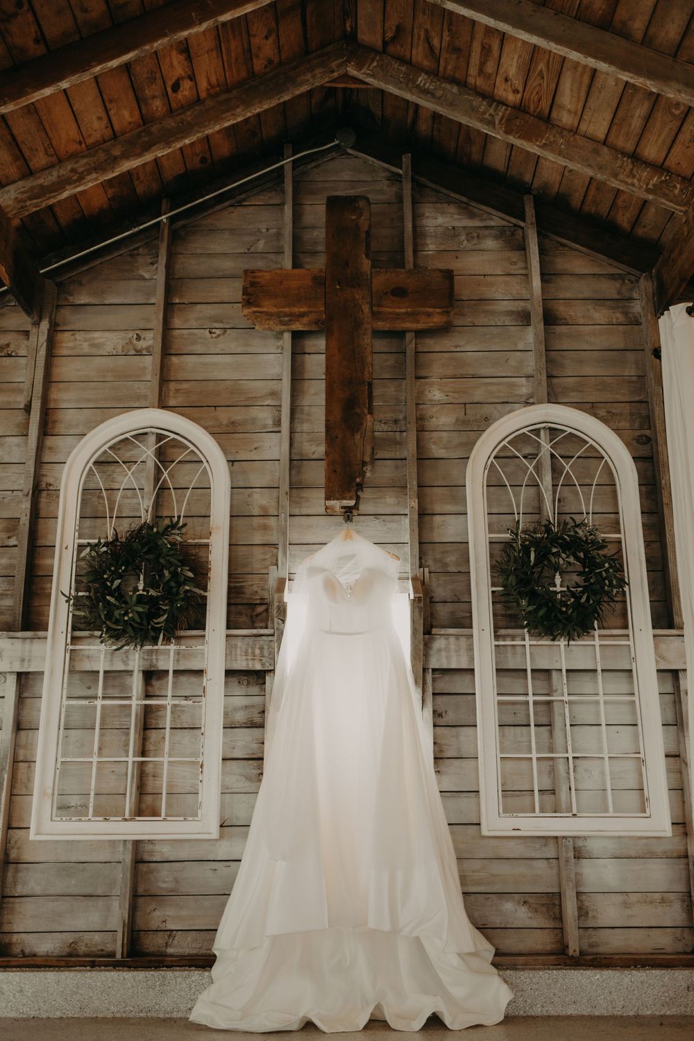 A white wedding dress hangs on rustic wooden church wall with arched windows and wooden cross.