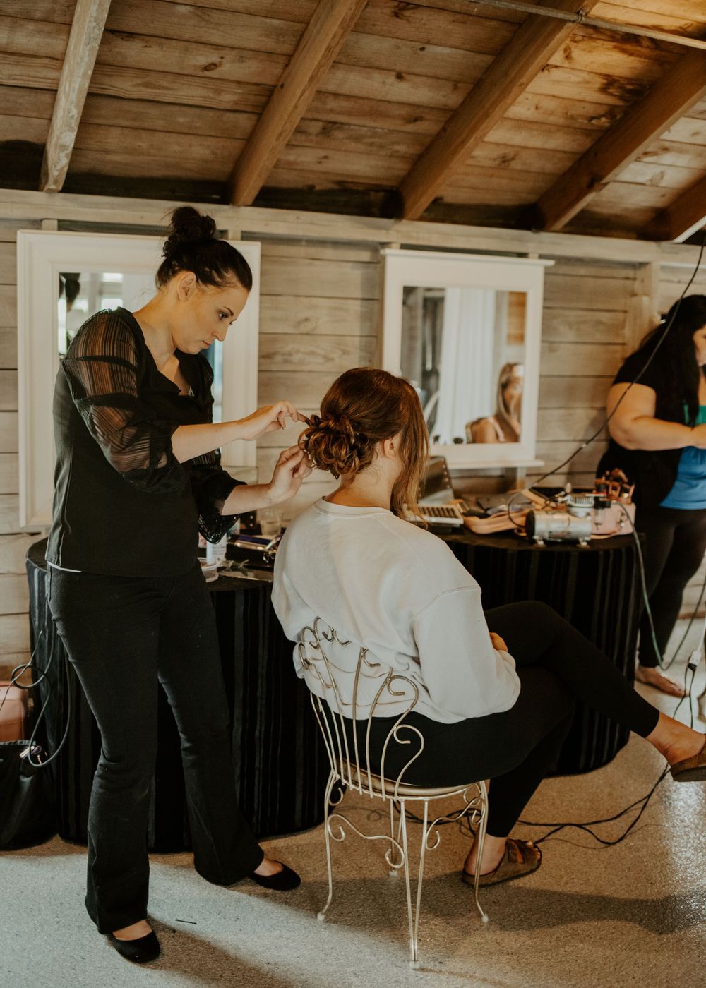 Behind the scenes wedding preparation showing stylist working on hair in rustic wooden room.