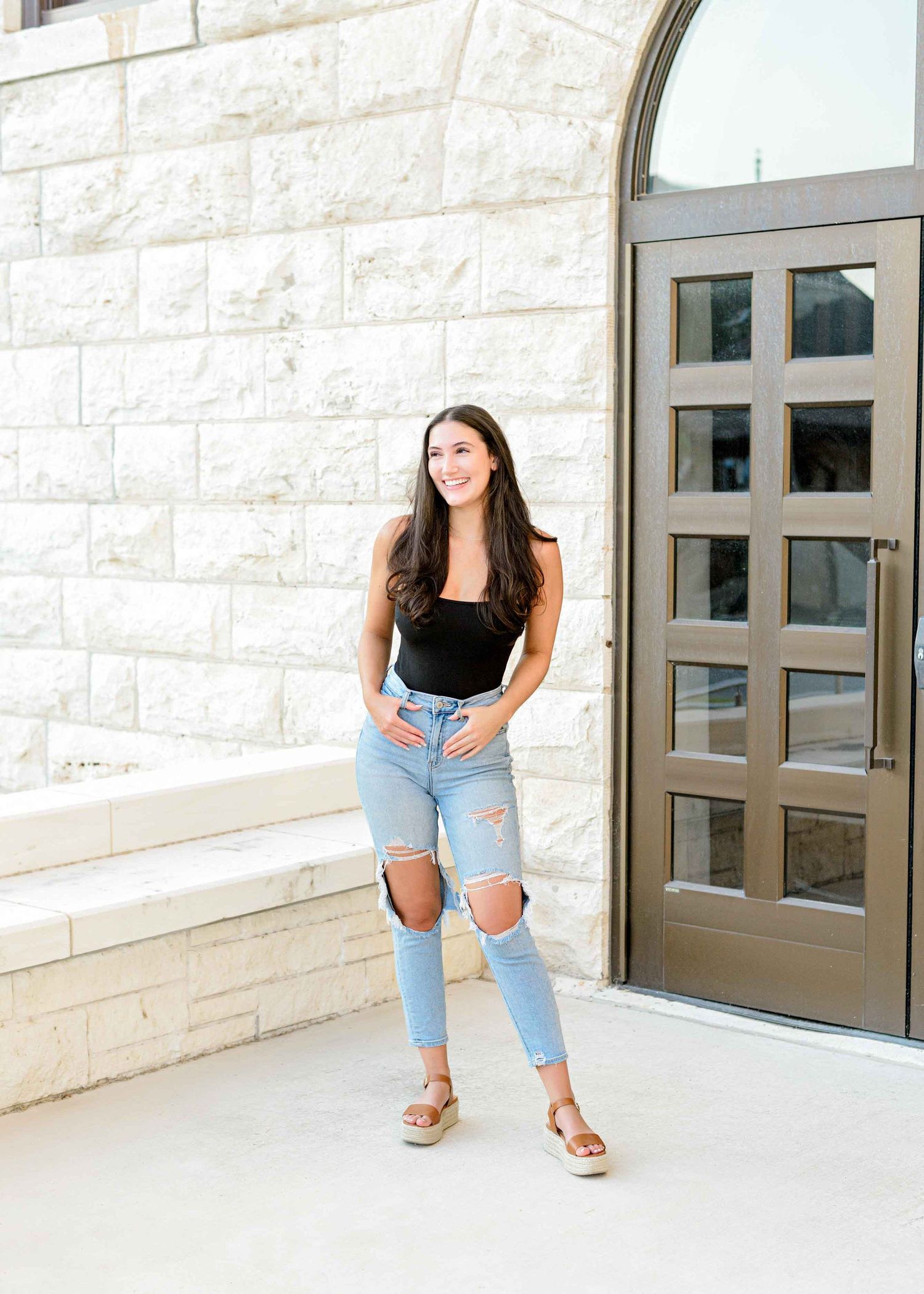 Casual portrait against white stone wall featuring distressed denim and black top with modern architecture.