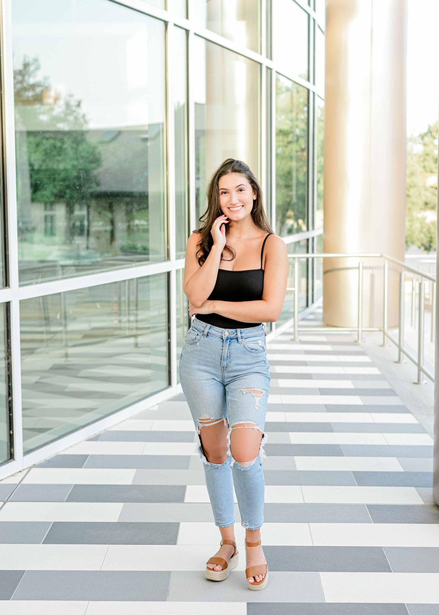 Casual lifestyle portrait in distressed jeans and black top along a modern corridor with glass walls.