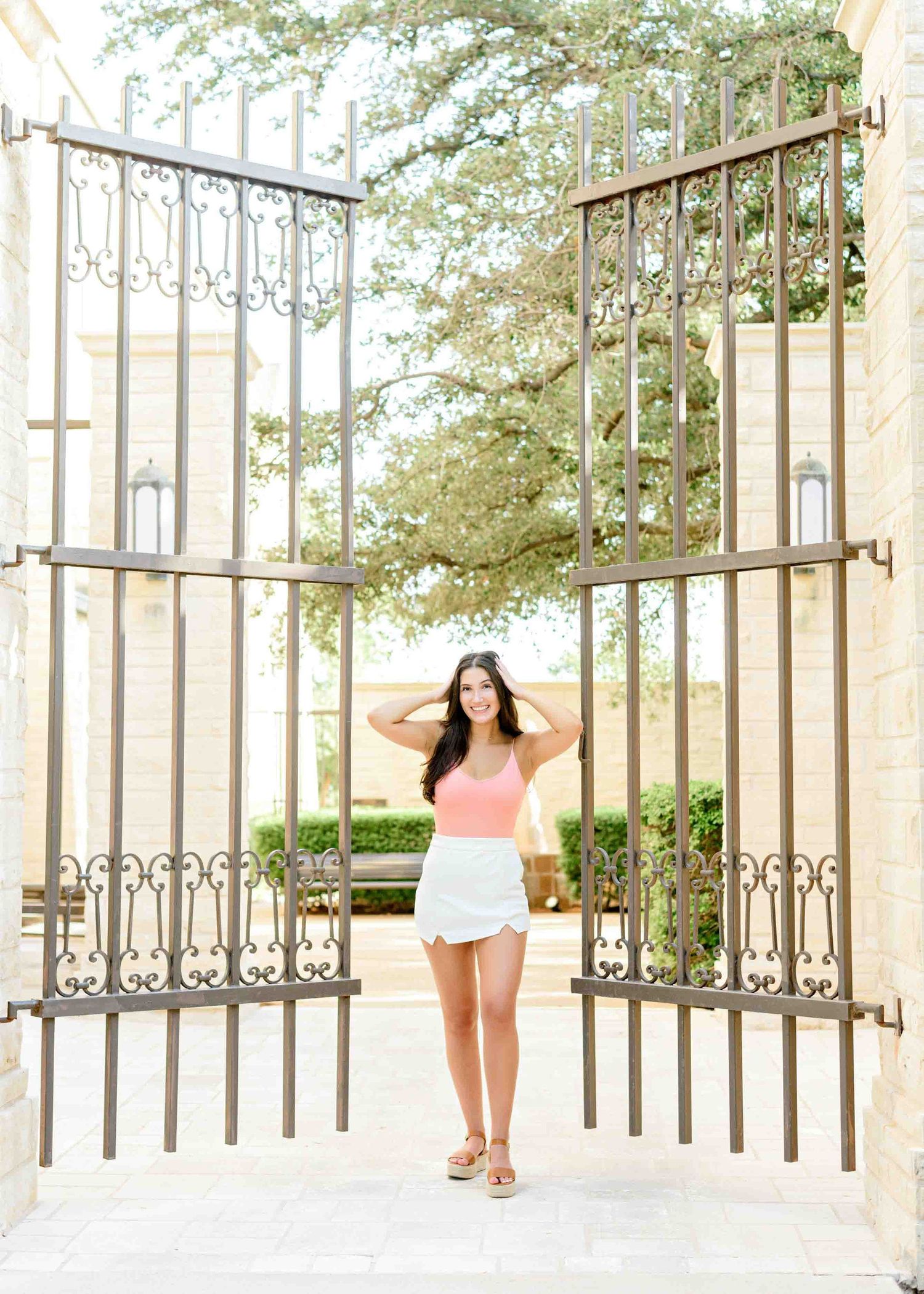Lifestyle portrait near ornate iron gates with stone pillars and greenery in background.