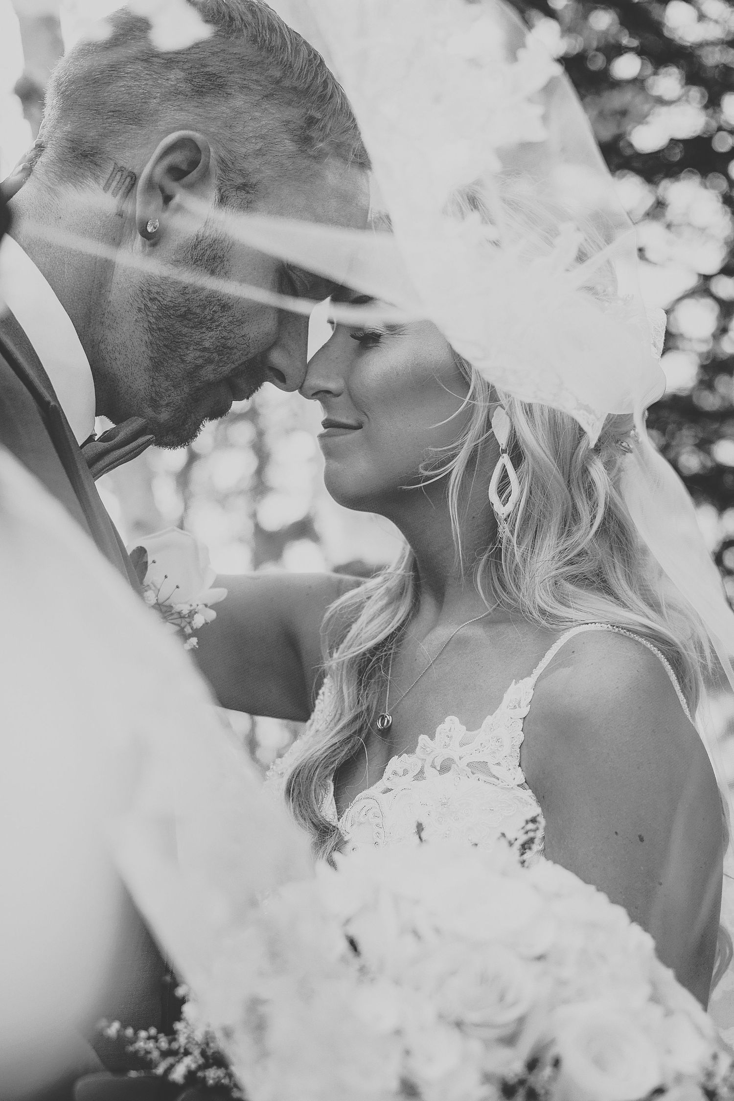 A romantic black and white wedding photograph showing a tender close-up moment with the bride in lace holding flowers.
