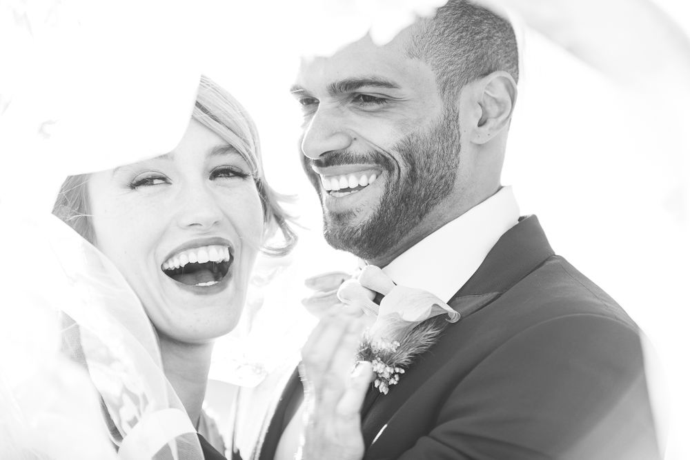 black and white photo of wedding couple under veil laughing