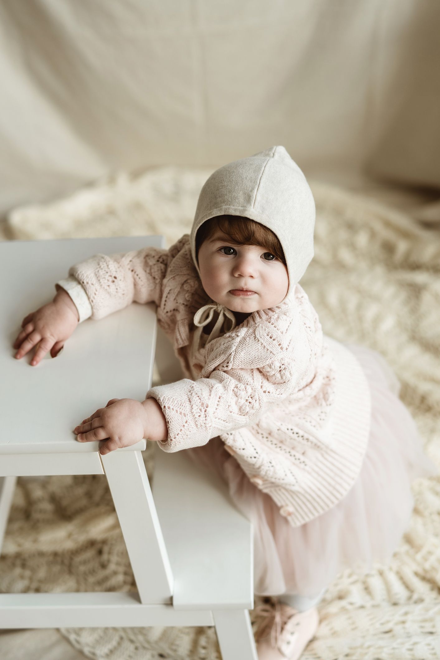 Child in bonnet and lace dress leaning on white chair against neutral backdrop.