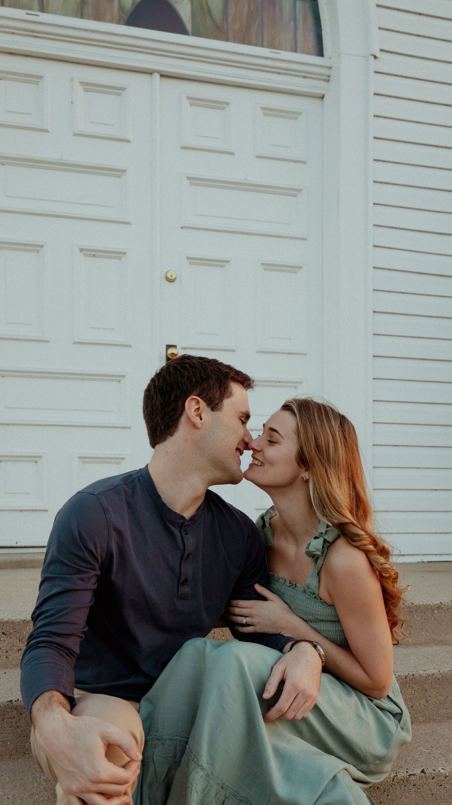 A romantic couple shares an intimate moment while sitting on steps in front of a white paneled door.