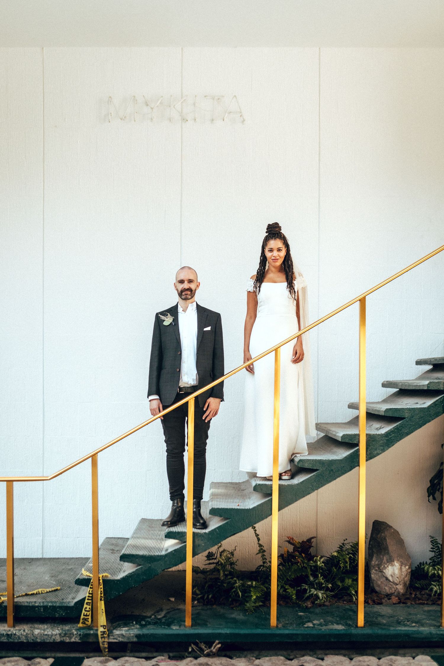 Wedding couple posing on green stairs with gold railings against a white wall.