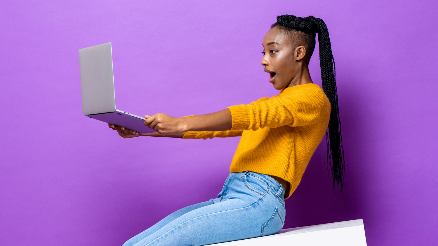Person in yellow sweater and jeans using laptop against purple background.