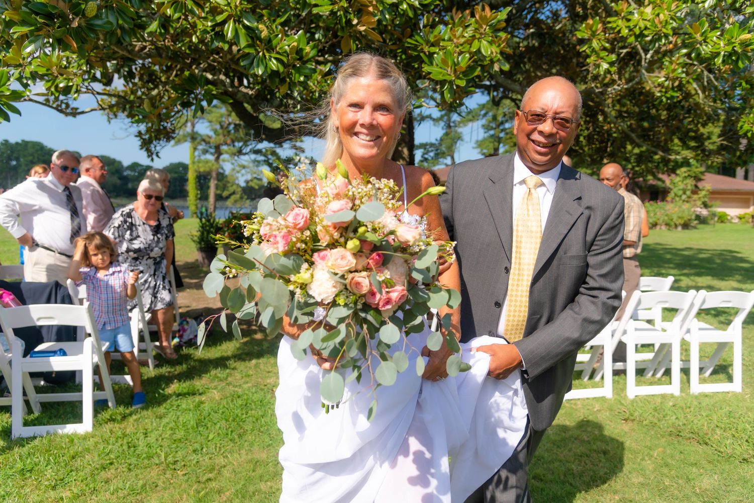 A joyful outdoor wedding ceremony with guests seated on white chairs under sunny skies.