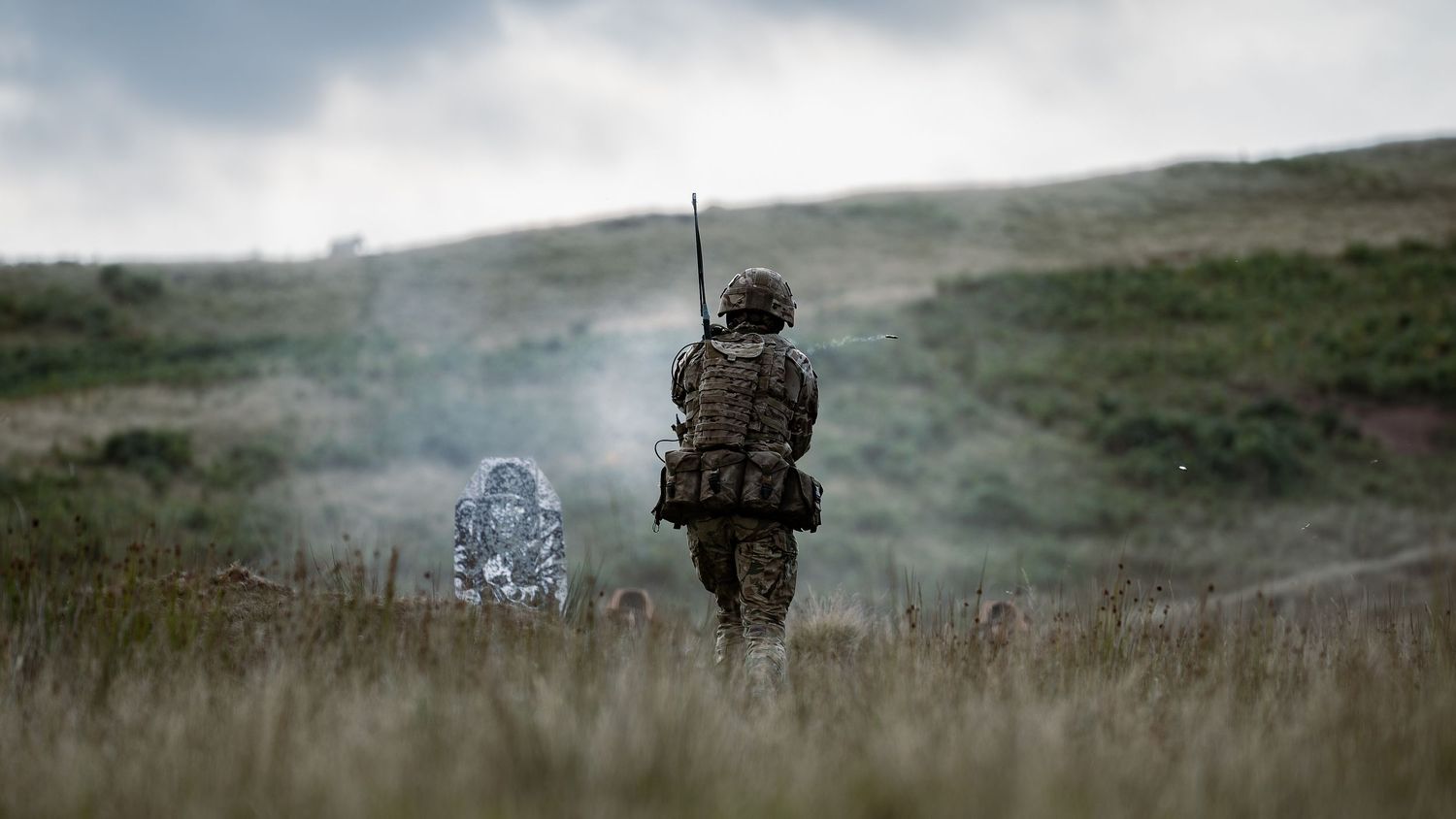Soldier walking through tall grass at dusk with equipment and gear.