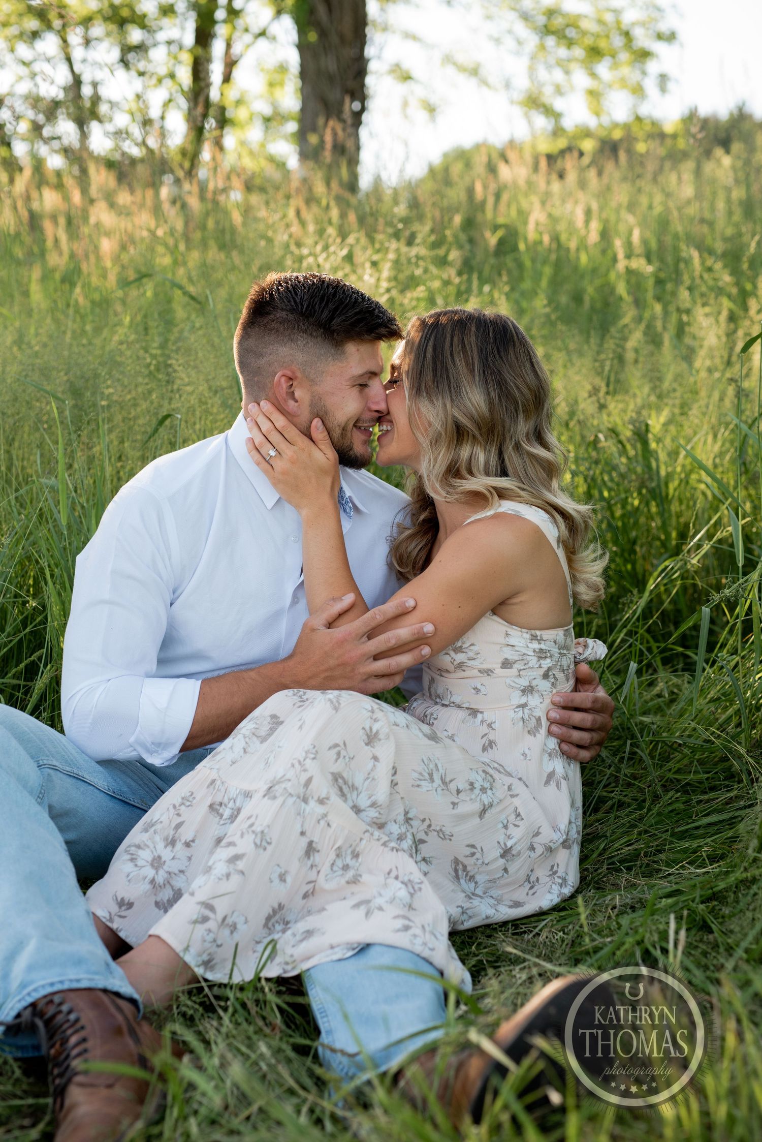 A romantic couple shares an intimate moment while sitting in tall grass during a sunny outdoor photoshoot.