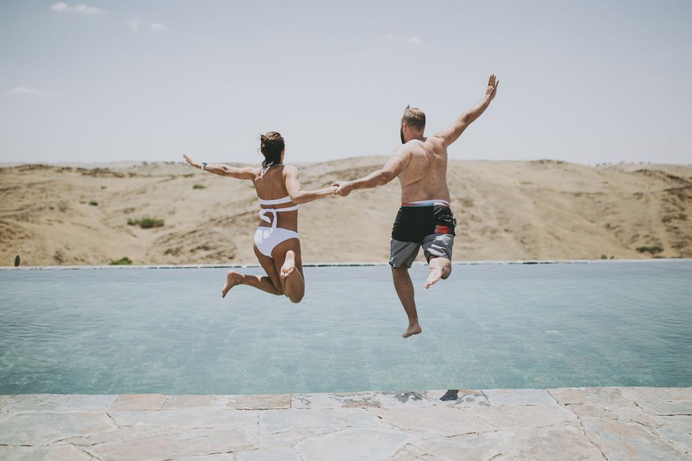Two people joyfully leap into bright blue water from a desert cliff edge.