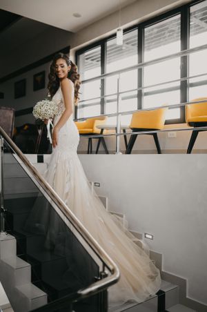 Bride in fitted lace gown poses on modern staircase with bouquet.