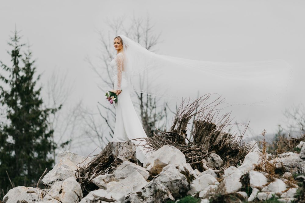 Bride in flowing white dress stands elegantly atop snow-covered rocks in winter landscape.