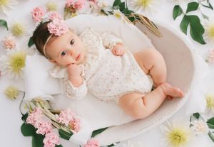 A baby in white lace outfit lies surrounded by pink and white flowers on white fabric.
