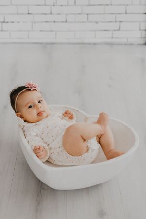 A baby wearing white lace clothing rests in a heart-shaped white bowl against a brick wall.