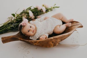 A baby in white lace outfit lying in a wooden bowl with flowers and greenery.
