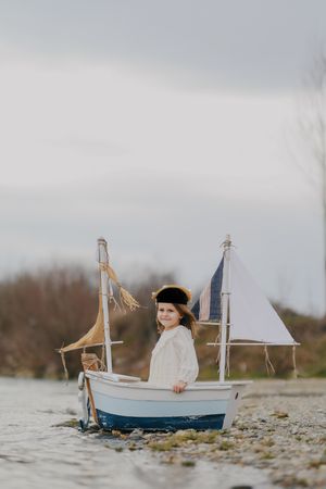 A small child sits in a blue and white toy sailboat on a rocky shore.