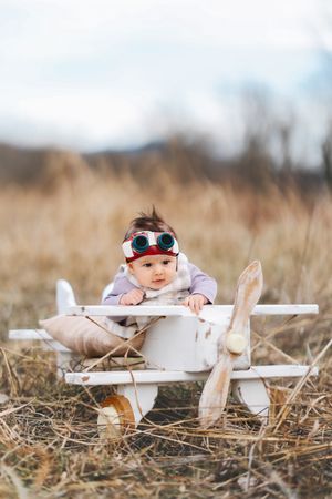 A baby wearing aviation gear sits in a decorative white wooden airplane in a field.