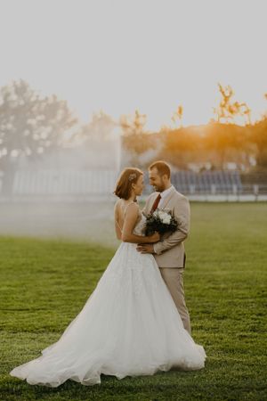 Newlyweds embracing on green lawn during sunset with bride in flowing white dress.