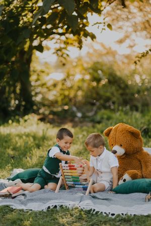 Two children playing with abacus on blanket outdoors with large teddy bear.