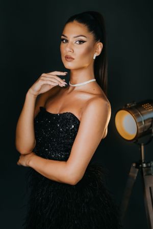Fashion model in black sequined dress poses against dark background with studio light.