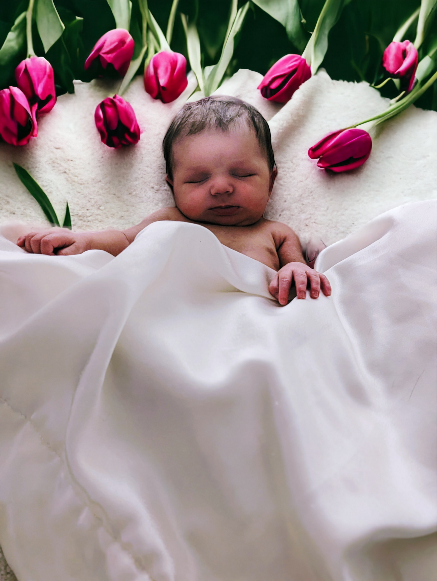 Newborn baby sleeping peacefully surrounded by pink tulips on white dreamy fabric.