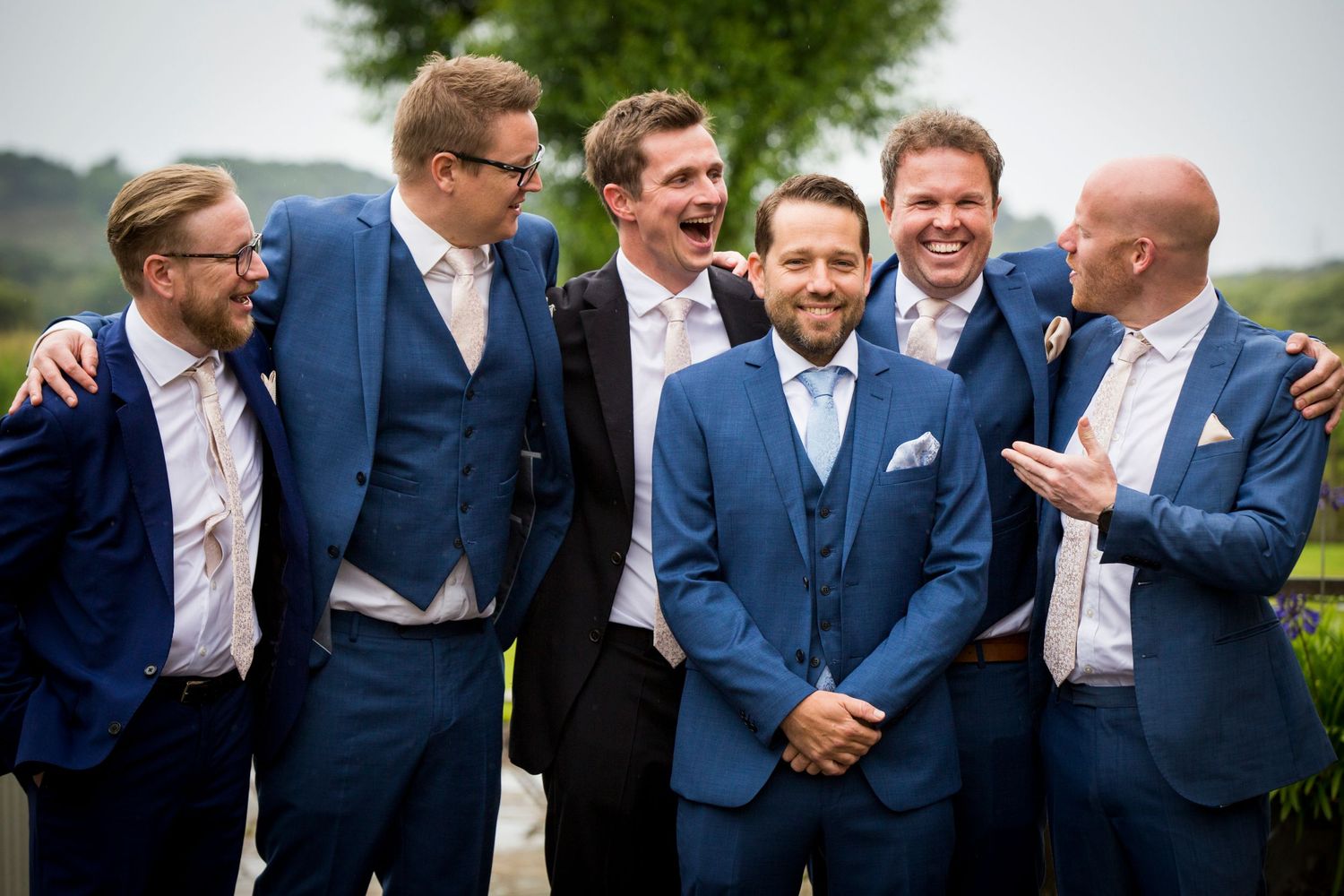 Groomsmen in blue suits share a lighthearted moment outdoors.