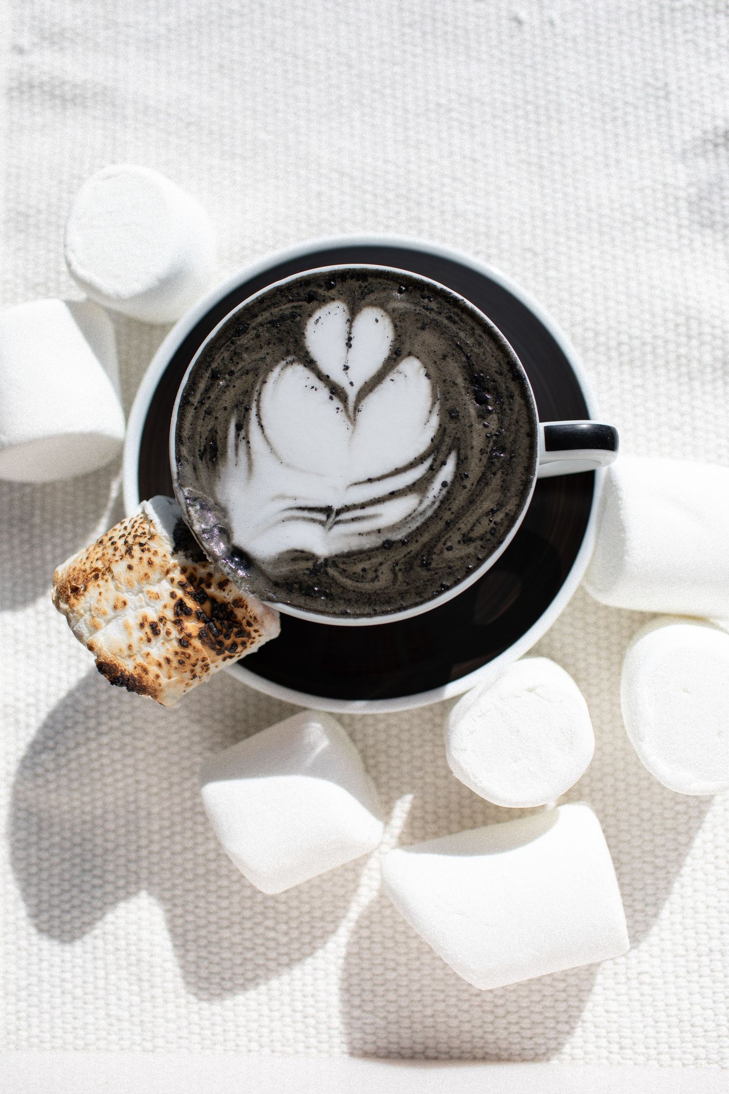 Artistic latte art designs in black coffee cups with marshmallows on white surface.