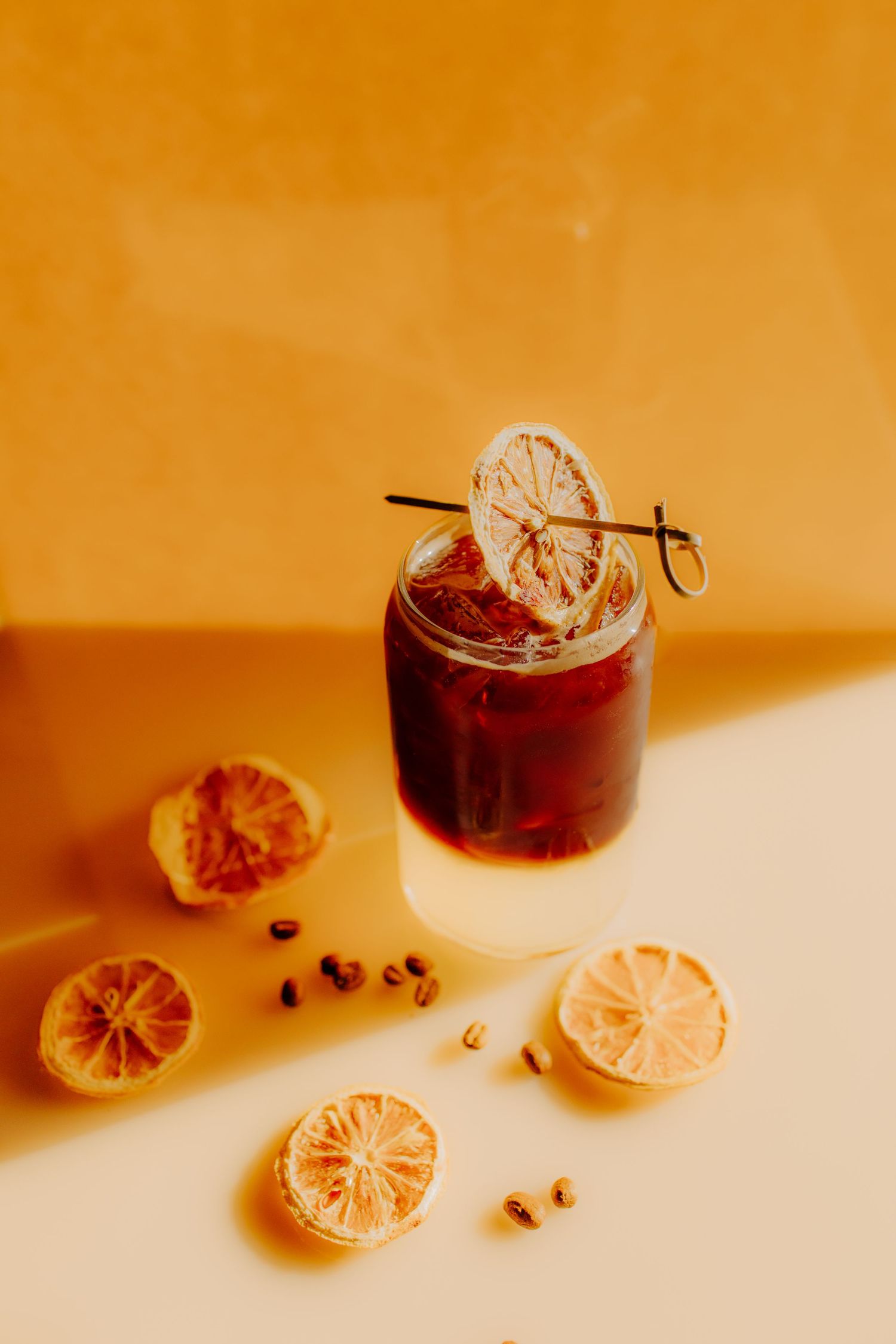 Orange citrus slices and dark beverage create a warm-toned minimalist composition against an amber background.