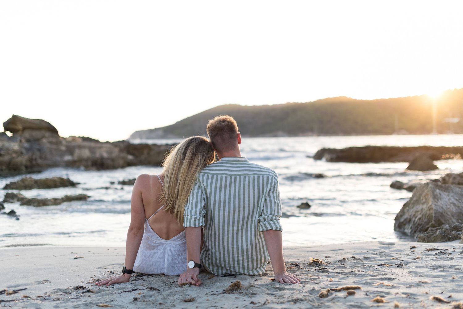 A couple sits on the sand as the woman leans on the mans shoulder and they watch the sun setting over the sea