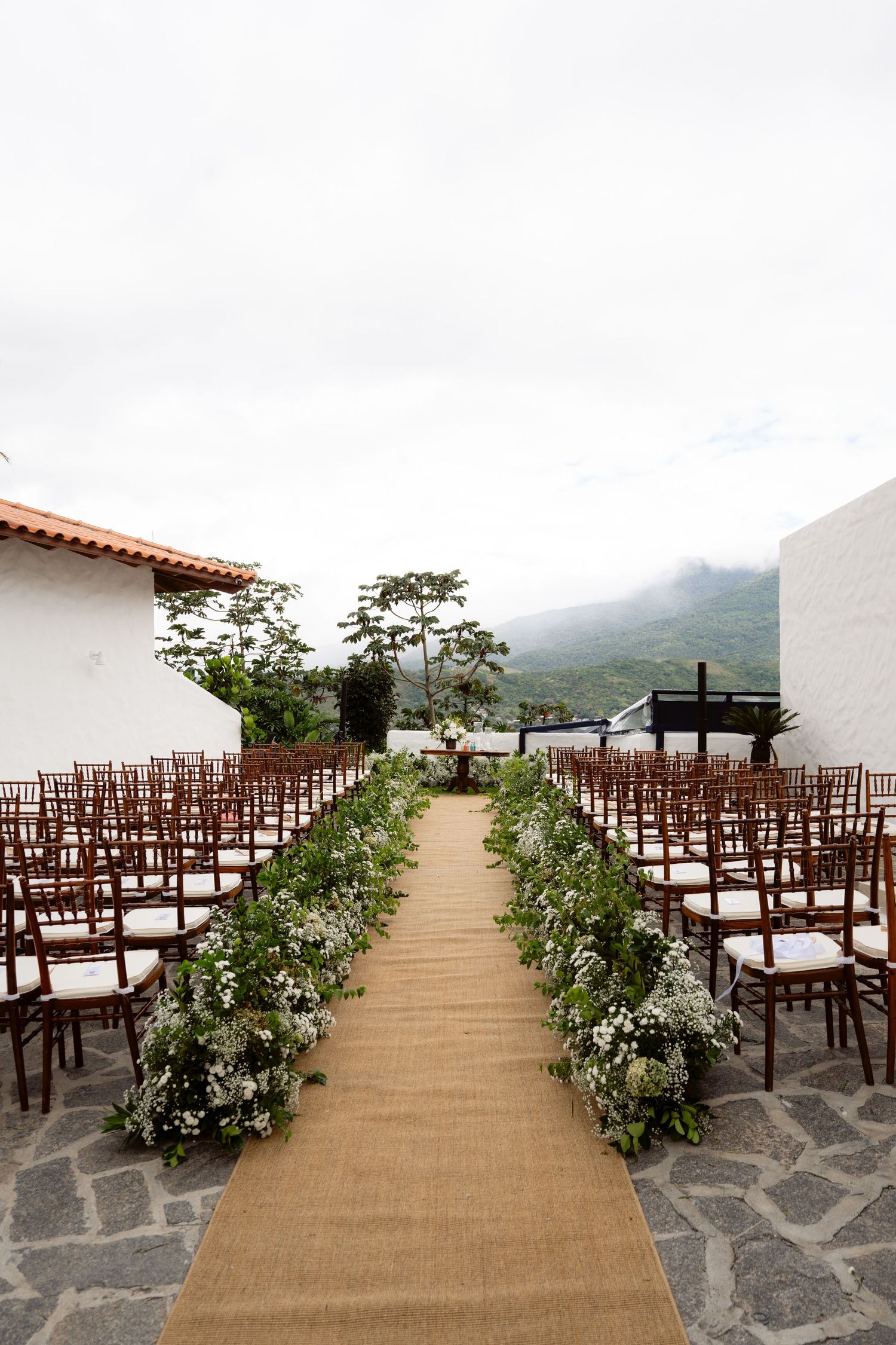 An elegant outdoor wedding ceremony setup with a burlap aisle runner lined with white floral arrangements.