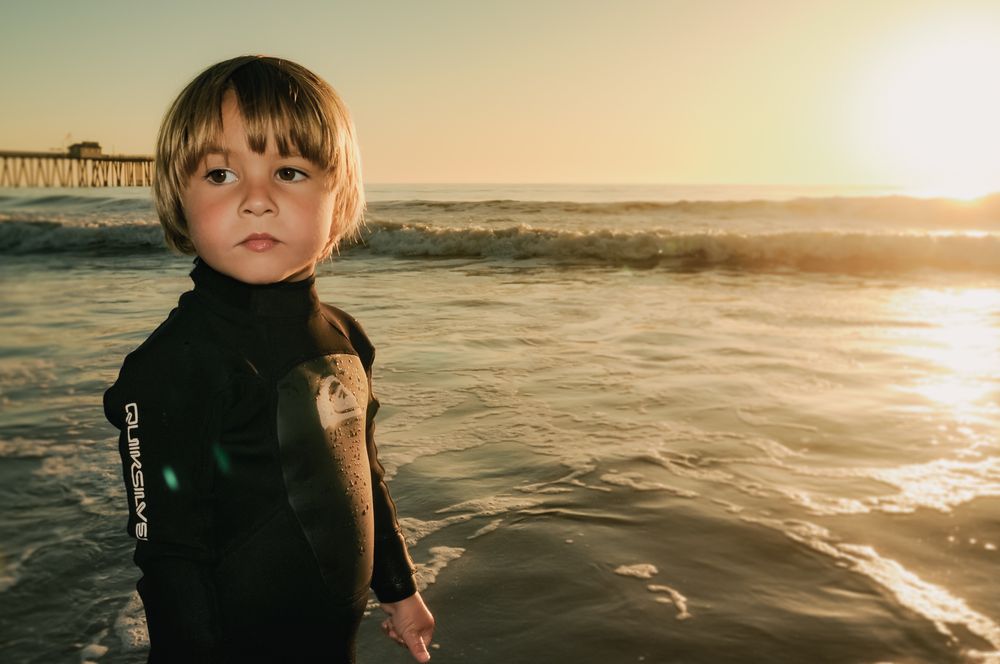 Young child in black wetsuit standing at ocean shore during golden sunset near pier.
