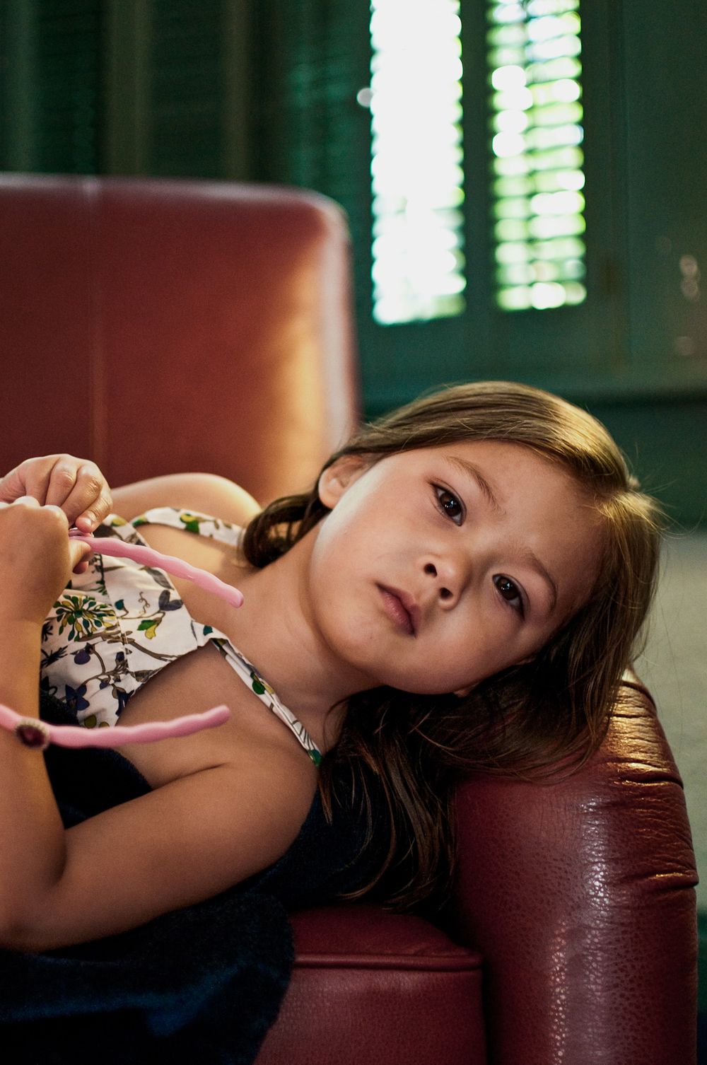 Young girl relaxing on red leather sofa holding pink toy near green shuttered window.