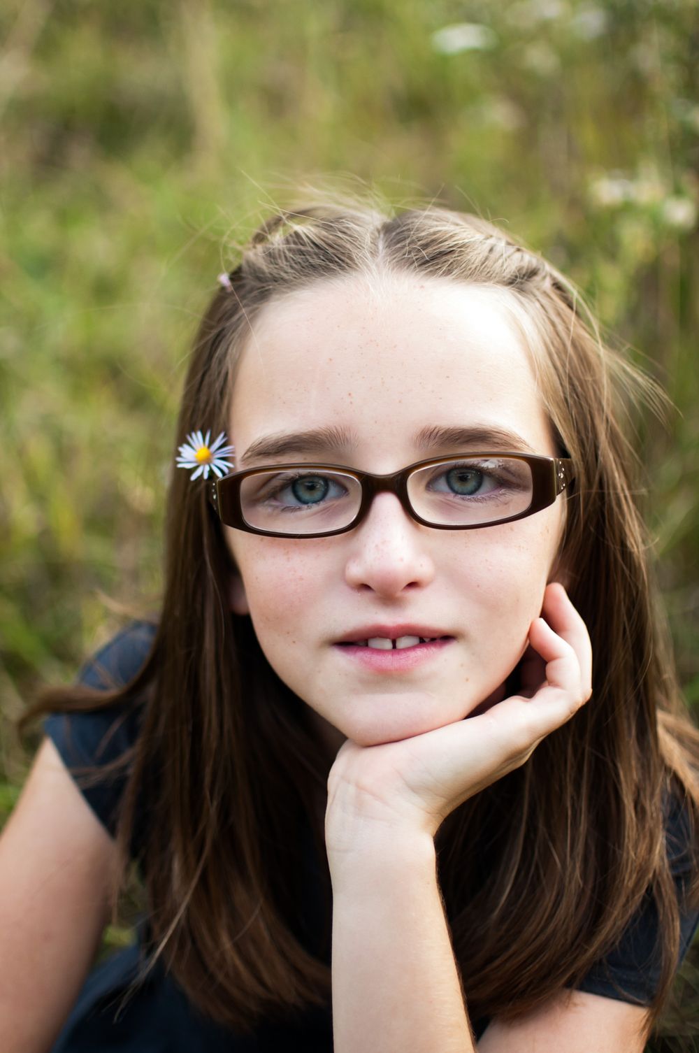 Preteen girl with glasses and daisy hair clip resting chin on hand in outdoor portrait.