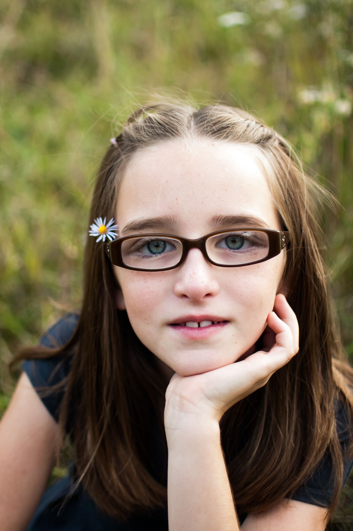 Preteen girl with glasses and daisy hair clip resting chin on hand in outdoor portrait.