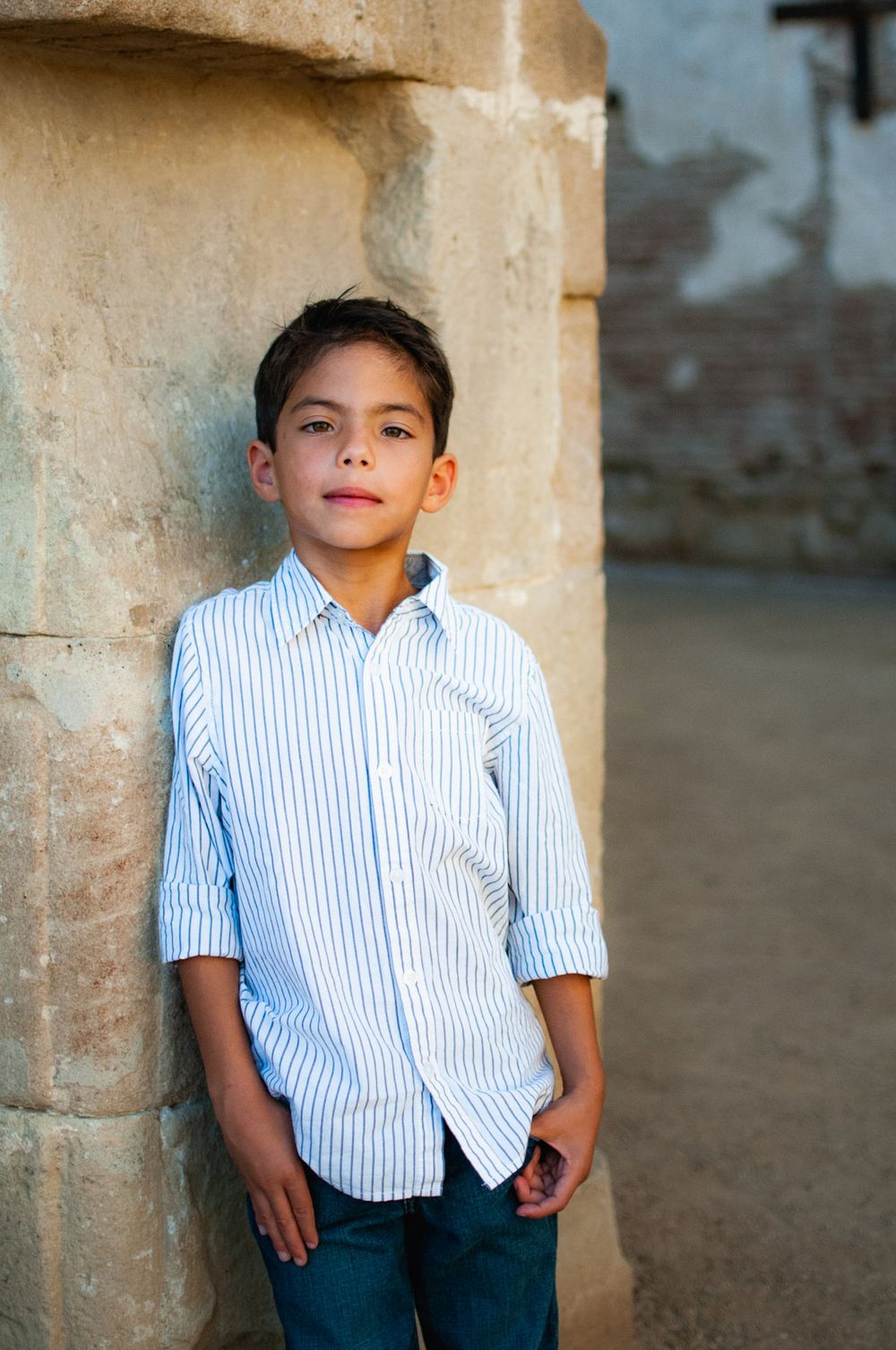 Young boy in striped blue shirt leaning against stone column outdoors in natural light.