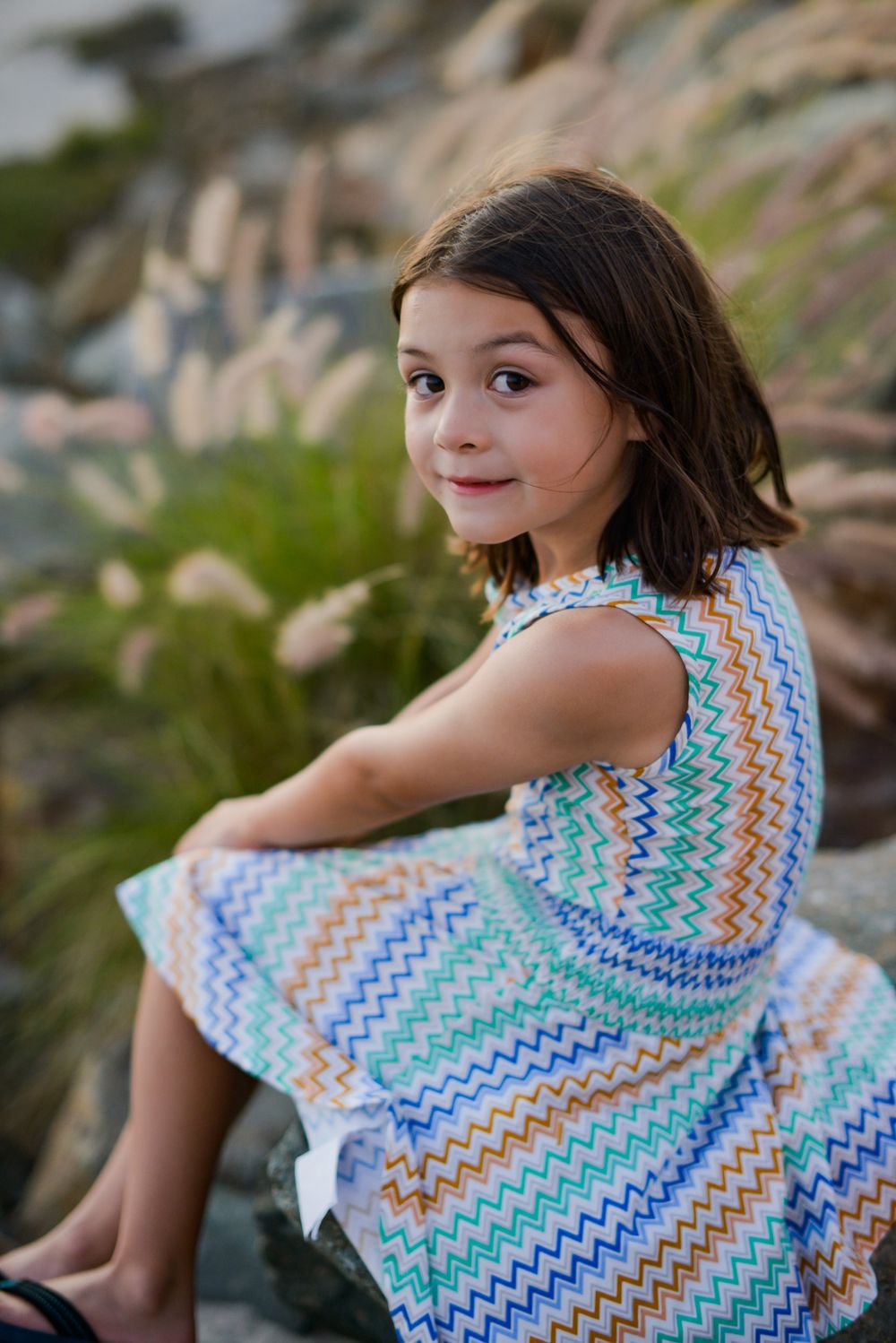 Young girl in colorful zigzag patterned dress sitting on rocks with plants in background.