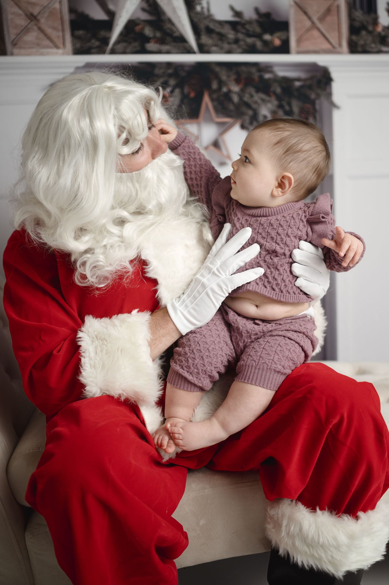 Santa Claus holds small baby wearing mauve knit outfit during Christmas photo session.