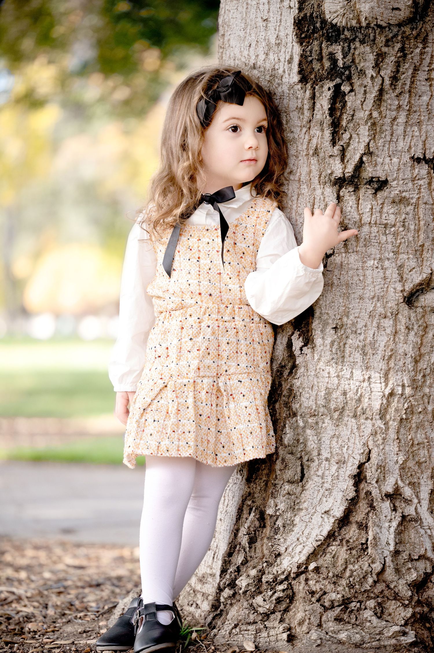 Young child in peach dress and white tights poses against large tree trunk.