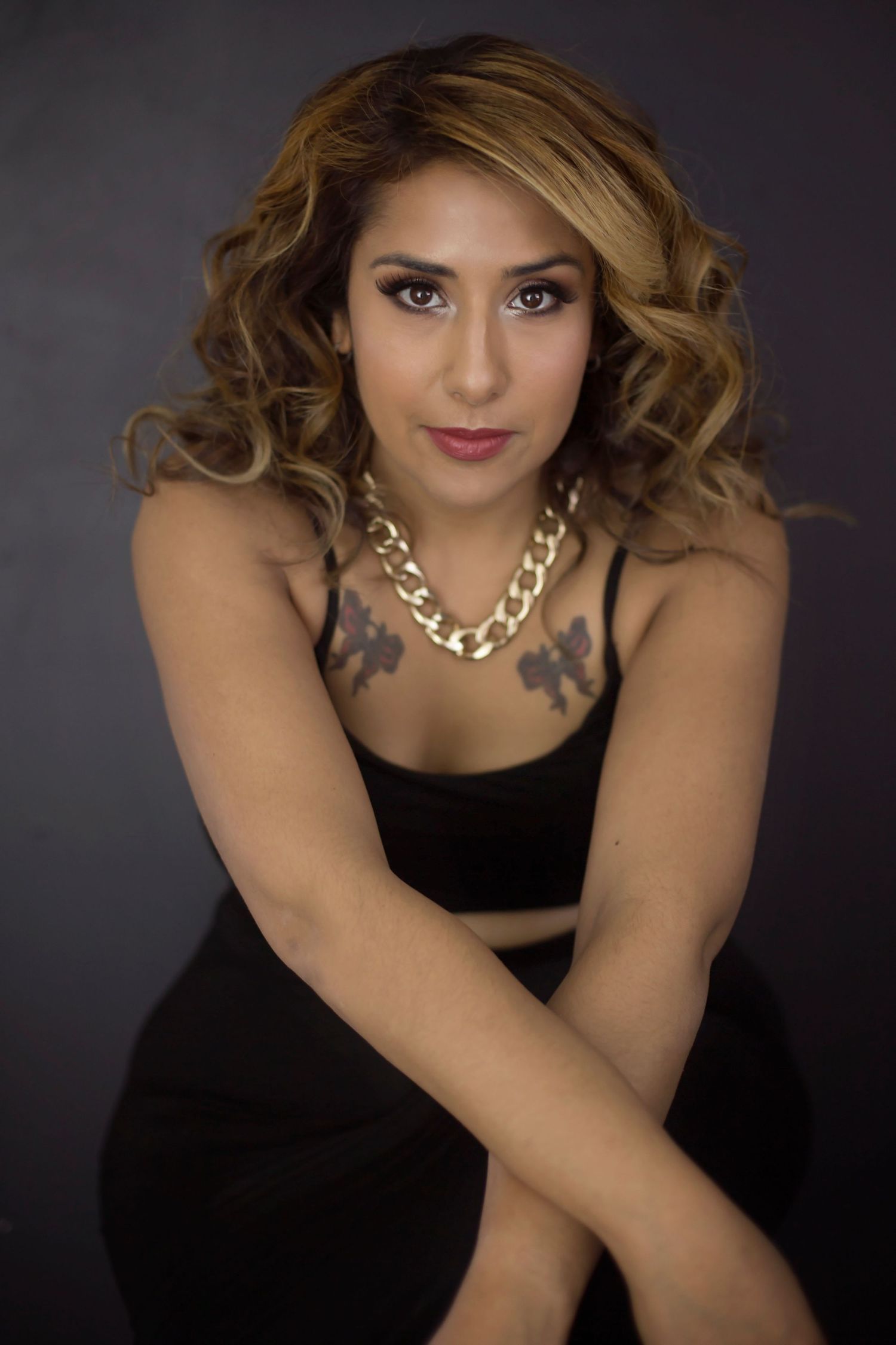 A woman with curly hair and chest tattoos wearing a black dress and gold necklace poses on a dark background.