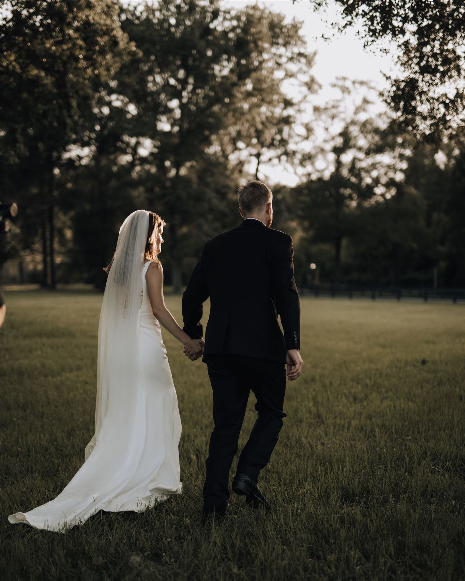 Newlyweds walk hand in hand across a sunlit grassy field during golden hour.